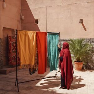 A woman in a red hijab is hanging clothes on a drying rack outdoors in a sunny courtyard. She adjusts various garments, including a hijab, illustrating how to wash hijabs and dry them with care.