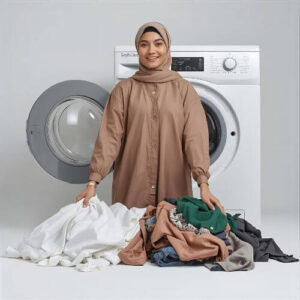 A woman in a beige dress stands in front of a washing machine, holding a pile of clothes, including a hijab. She is preparing to wash her garments, demonstrating how to wash hijabs along with other laundry items.