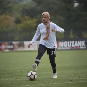 A female athlete wearing a beige hijab while playing football. Her hijab is an example of what is a hijab worn in active sports, promoting modesty and practicality. The hoor hijab n abayas collection can also include sporty, comfortable options like this