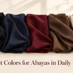 Hoor Hijab & Abayas flat lay showing five folded abaya fabric swatches in black, navy, deep maroon, coffee brown, and beige arranged in a gradient row on a warm neutral background with soft natural lighting and elegant fashion styling.